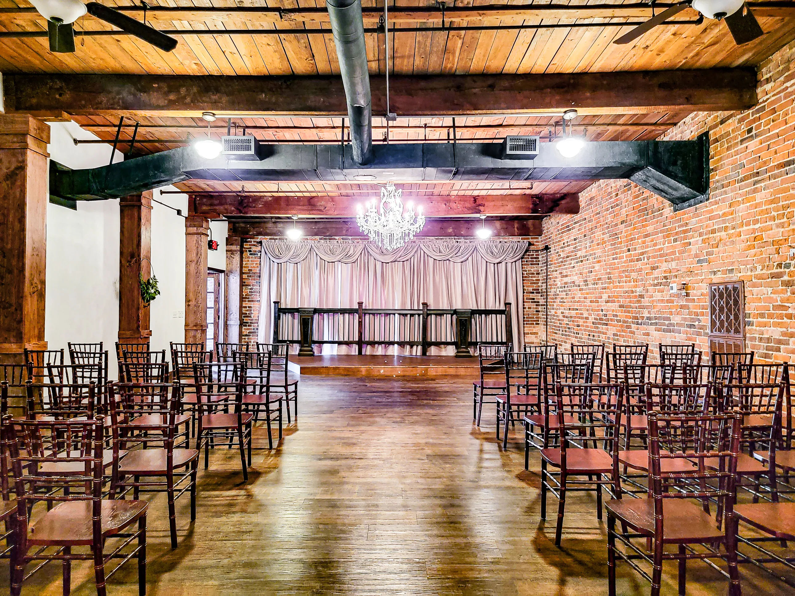Ceremony chairs facing a draped backdrop beneath timber beams and chandeliers at The Cotton Loft
